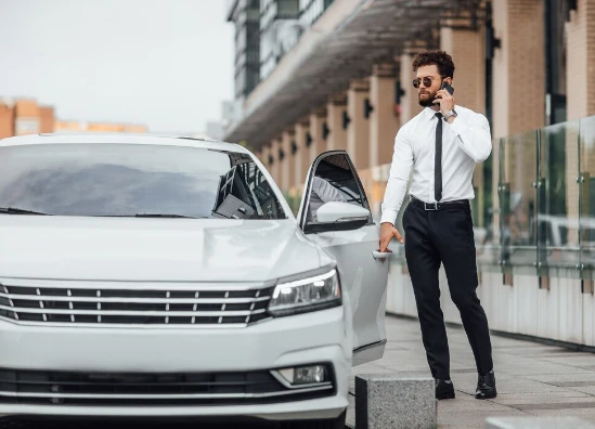 Homme en chemise blanche et cravate au téléphone près de la portière ouverte d’une voiture blanche.